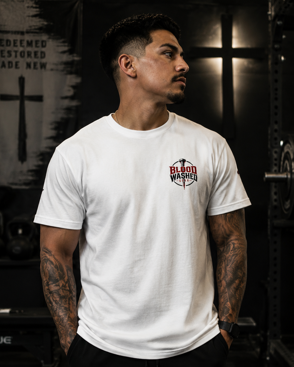 A man with tattoos wearing a white crewneck t-shirt with a red and black Christian Apparel 'Blood Wahed' logo on the left chest, standing in a dark gym environment.